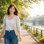 朗らかな朝の光の中、川沿いの遊歩道を笑顔で散歩する大人の女性。白Tとベージュのカーディガン、ハイウエストデニムにスニーカーのカジュアルコーデで、木漏れ日と柔らかなボケが心地よい横長ポートレート。 A cheerful adult woman walking along a riverside path in soft morning light, smiling warmly in a casual outfit (white tee, beige cardigan, high-waist jeans, sneakers) with gentle bokeh and fresh greenery in a wide landscape portrait.